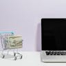 Mini shopping cart with cash beside a laptop, symbolizing online shopping and e-commerce. Ebay